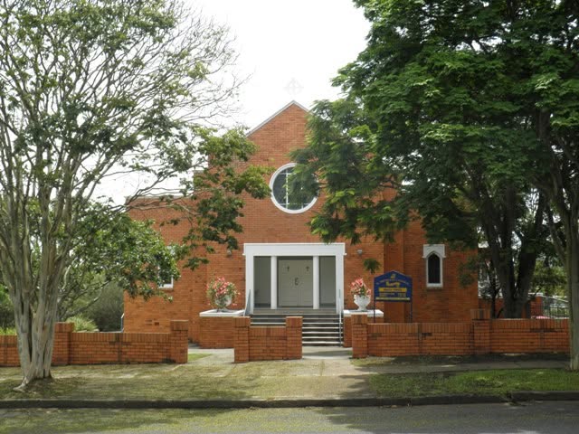 A brick church building with a circular window, surrounded by green trees and a small garden. The entrance features steps leading to double doors, and colorful flowers are placed beside the entrance.