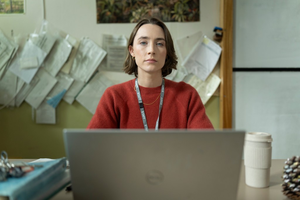 A young woman sitting at a desk, looking directly at the camera, with a laptop in front of her and papers pinned on the wall behind her.