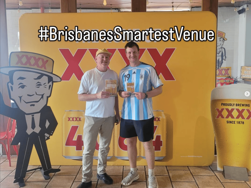 Two men posing in front of a bright yellow backdrop featuring the XXXX beer brand, holding promotional cards. One man wears a white shirt and the other is in an Argentina soccer jersey. The hashtag #BrisbanesSmartestVenue is displayed above them.
