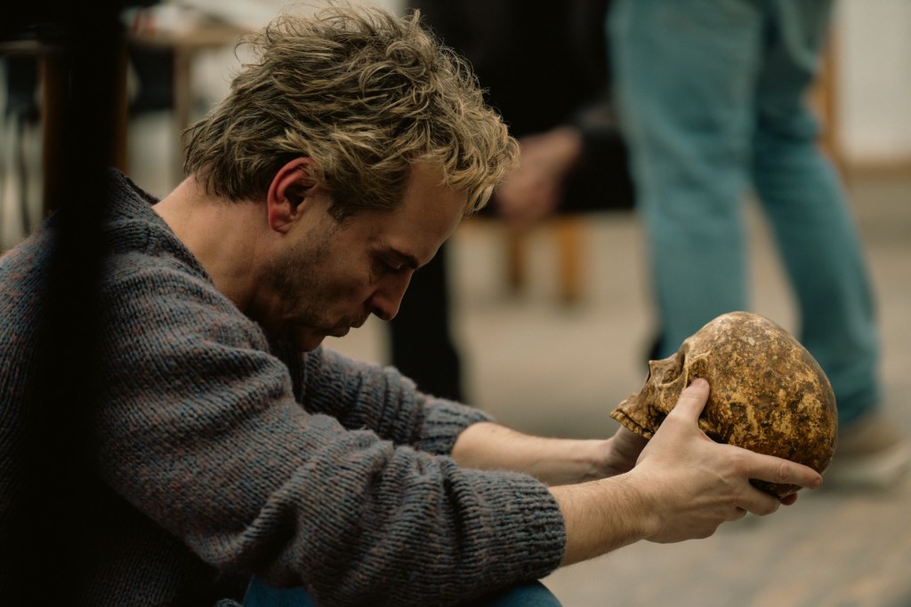 A man sitting on the floor, holding a skull in his hands, with a contemplative expression.