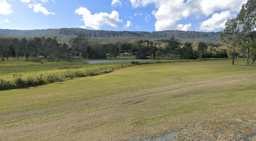 A scenic view of a grassy field leading to a winding road, surrounded by trees and mountains under a partly cloudy sky.