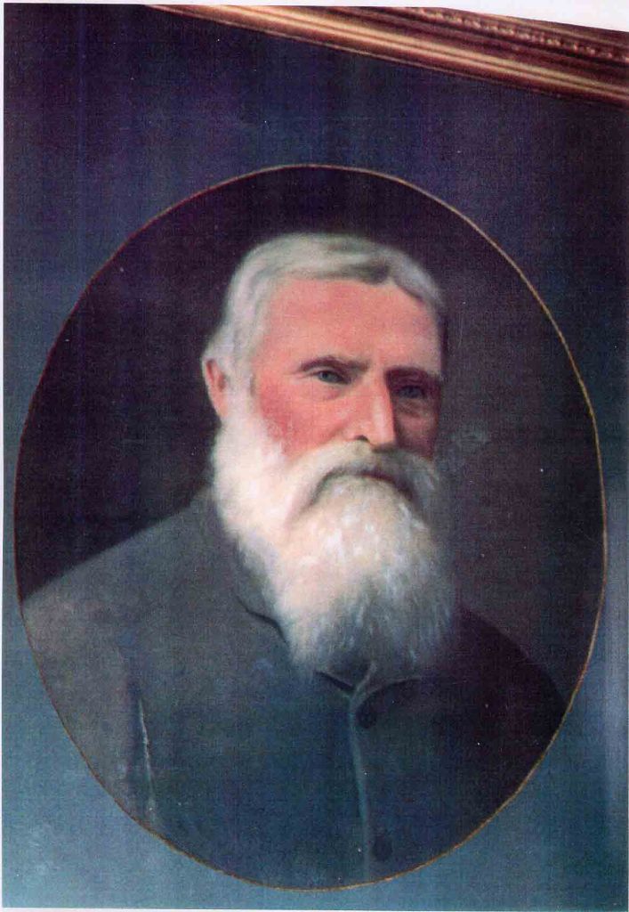 Portrait of an elderly man with a white beard, wearing a gray jacket, set against a dark background.
