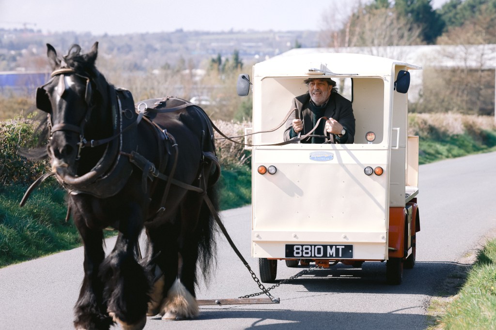 A man with a hat smiles while driving a small, vintage-style cart being pulled by a horse along a rural road surrounded by greenery.